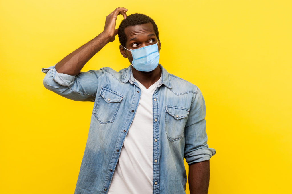 Portrait of uncertain young man with surgical medical mask looking up, scratching head while thinking with confused puzzled face.